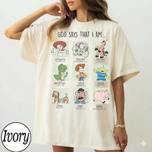 Puede incluir: Camiseta color crema con el texto "GOD SAYS THAT I AM..." e ilustraciones de personajes de dibujos animados. Cada personaje está etiquetado con una afirmación positiva y un versículo bíblico.