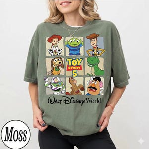 Pode incluir: Camiseta verde-oliva com uma grade de personagens de Toy Story, incluindo Woody, Buzz Lightyear e Rex. A camisa tamb&eacute;m tem o texto "Toy Story 5" e "Walt Disney World" em preto e amarelo.