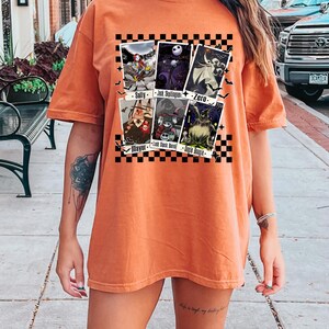 May include: Orange t-shirt featuring a graphic of characters from The Nightmare Before Christmas. The design includes images of Sally, Jack Skellington, Zero, Mayor, Lock, Shock, Barrel, and Oogie Boogie. The design is set against a black and white checkered background.