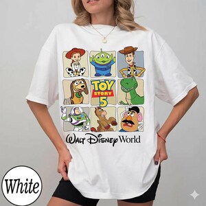 Pode incluir: Camiseta branca com uma grade de personagens de Toy Story, incluindo Woody e Buzz Lightyear. A camiseta tamb&eacute;m tem o texto "Toy Story 5" e "Walt Disney World".