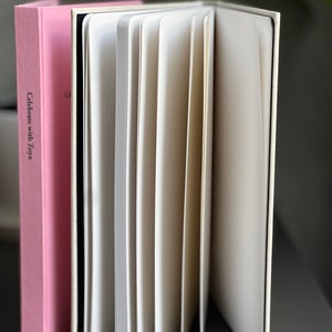 Op de afbeelding: Een open notitieboek met gebroken witte pagina's en een harde kaft, naast een roze boek met de tekst "Celebrate with Zoya". De pagina's van het notitieboek zijn uitgespreid en onthullen hun lege binnenkant. De boeken staan op een donkere ondergrond.