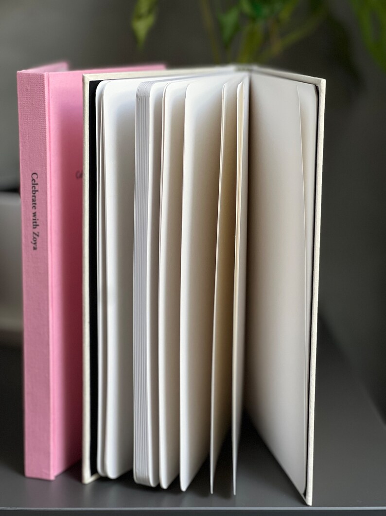 Op de afbeelding: Een open notitieboek met gebroken witte pagina's en een harde kaft, naast een roze boek met de tekst "Celebrate with Zoya". De pagina's van het notitieboek zijn uitgespreid en onthullen hun lege binnenkant. De boeken staan op een donkere ondergrond.