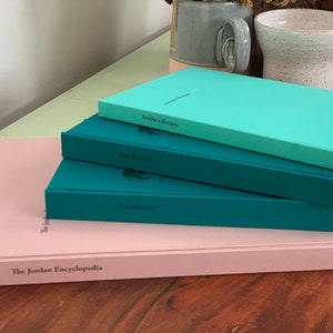 Op de afbeelding: Een stapel hardcover boeken in verschillende kleuren: lichtroze, blauwgroen en turkoois. De titels zijn onder meer "The Jordan Encyclopedia", "Max Ryerson" en "Annie's Recipes". De boeken liggen op een houten oppervlak, met een lichtgroene tafel en keramische mokken op de achtergrond.