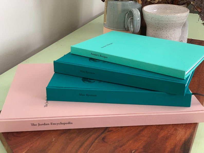 Op de afbeelding: Een stapel hardcover boeken in verschillende kleuren: lichtroze, blauwgroen en turkoois. De titels zijn onder meer "The Jordan Encyclopedia", "Max Ryerson" en "Annie's Recipes". De boeken liggen op een houten oppervlak, met een lichtgroene tafel en keramische mokken op de achtergrond.
