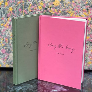 Op de afbeelding: Twee hardcover journals, &eacute;&eacute;n saliegroen en &eacute;&eacute;n roze, worden getoond. Het roze journal heeft de tekst "slay the day" en "KIM SERA" in zwart script. Het groene journal heeft de tekst "slay the day" en "SHAVE SOME" in zwart script.