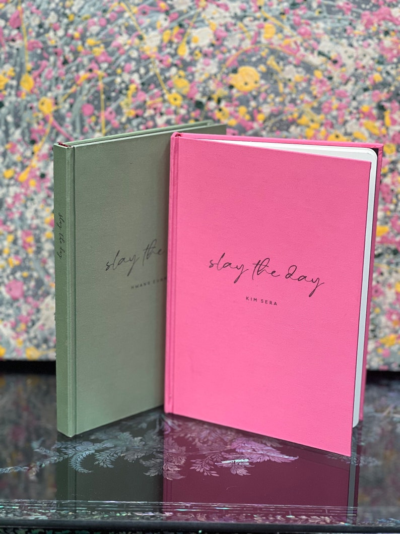 Op de afbeelding: Twee hardcover journals, &eacute;&eacute;n saliegroen en &eacute;&eacute;n roze, worden getoond. Het roze journal heeft de tekst "slay the day" en "KIM SERA" in zwart script. Het groene journal heeft de tekst "slay the day" en "SHAVE SOME" in zwart script.