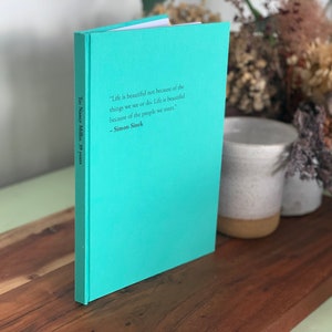 Op de afbeelding: Een hardcover boek met een turquoise omslag staat op een houten oppervlak. Het boek bevat de tekst: "Het leven is niet mooi vanwege de dingen die we zien of doen. Het leven is mooi vanwege de mensen die we ontmoeten." - Simon Sinek. De rug leest "To Nancy Miller, 39 years."