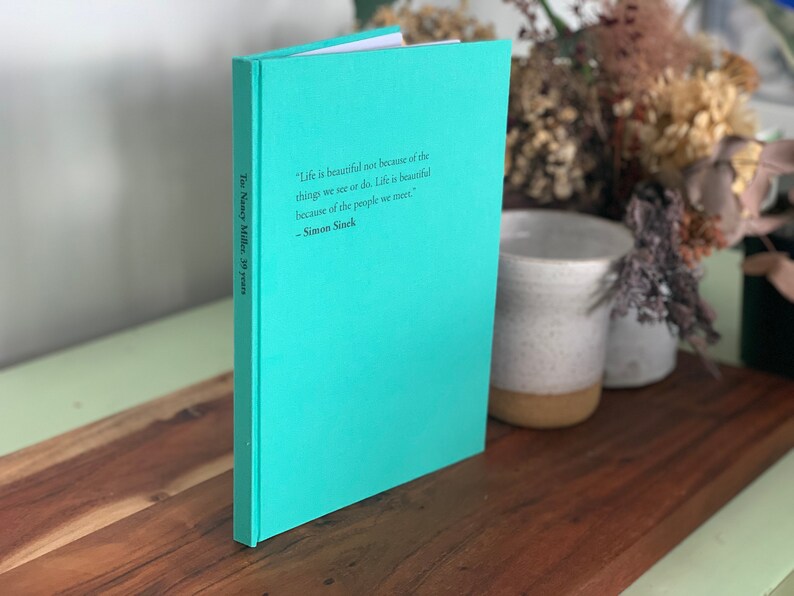 Op de afbeelding: Een hardcover boek met een turquoise omslag staat op een houten oppervlak. Het boek bevat de tekst: "Het leven is niet mooi vanwege de dingen die we zien of doen. Het leven is mooi vanwege de mensen die we ontmoeten." - Simon Sinek. De rug leest "To Nancy Miller, 39 years."