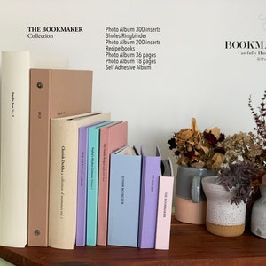 Op de afbeelding: Een verzameling fotoalbums en receptenboeken in pastelkleuren, waaronder cr&egrave;me, beige, lavendel, roze en blauw. De boeken zijn gerangschikt op een houten oppervlak, met de tekst "THE BOOKMAKER" zichtbaar. Droogbloemen in vazen staan op de achtergrond.