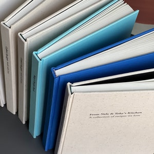 Op de afbeelding: Een verzameling kookboeken met verschillende gekleurde kaften, waaronder wit, lichtblauw en donkerblauw. De tekst "From Nele & Toby's Kitchen" is zichtbaar op &eacute;&eacute;n boek. De boeken zijn diagonaal gerangschikt.