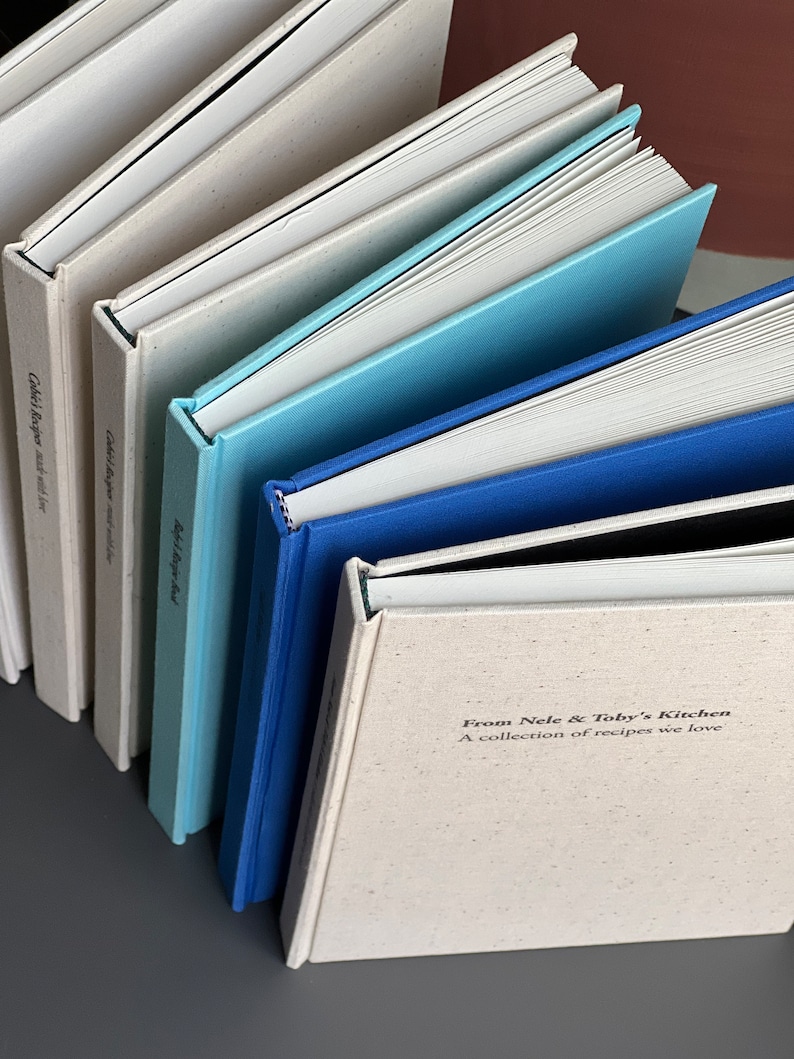 Op de afbeelding: Een verzameling kookboeken met verschillende gekleurde kaften, waaronder wit, lichtblauw en donkerblauw. De tekst "From Nele & Toby's Kitchen" is zichtbaar op &eacute;&eacute;n boek. De boeken zijn diagonaal gerangschikt.