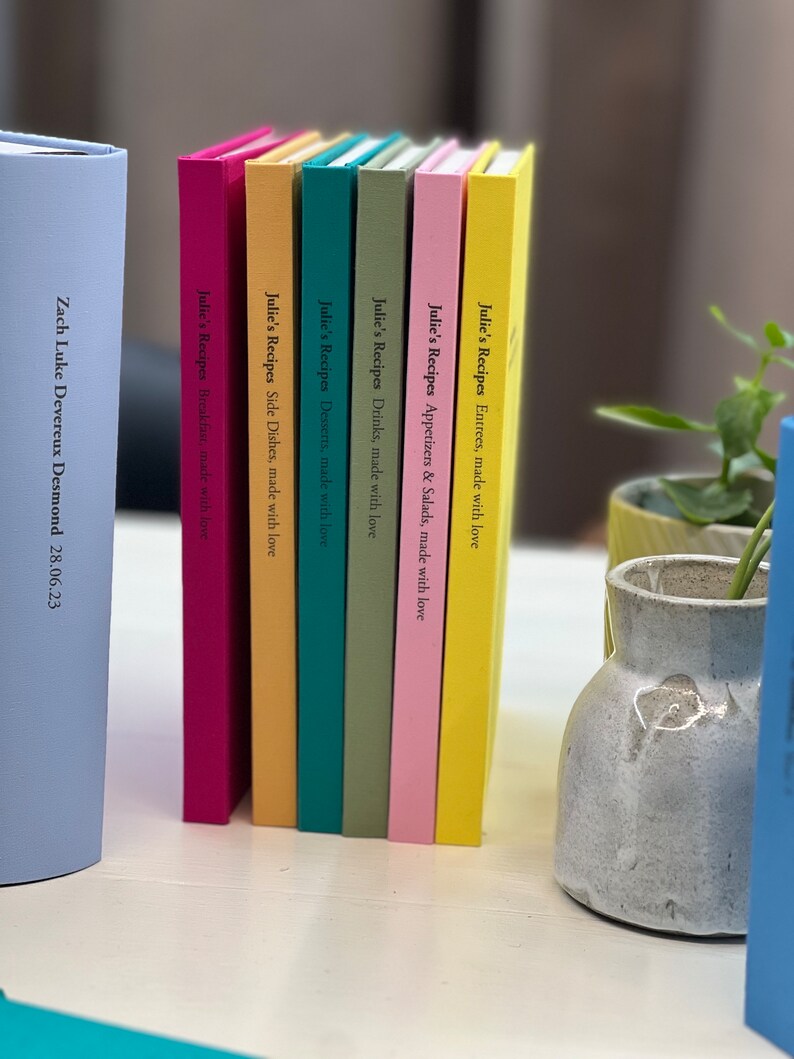 Op de afbeelding: Een verzameling kleurrijke receptenboeken met de tekst "Julie's Recipes" en verschillende maaltijdcategorie&euml;n. De boeken zijn in tinten roze, geel, groen en blauwgroen. Links staat een lichtblauw boek met tekst.
