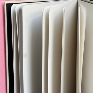 Op de afbeelding: Een open notitieboek met witte pagina's, dat het interieur onthult. De pagina's zijn uitgespreid, wat hun dikte en textuur laat zien. Links is een roze boek zichtbaar en een zwarte rug is zichtbaar achter de open pagina's.