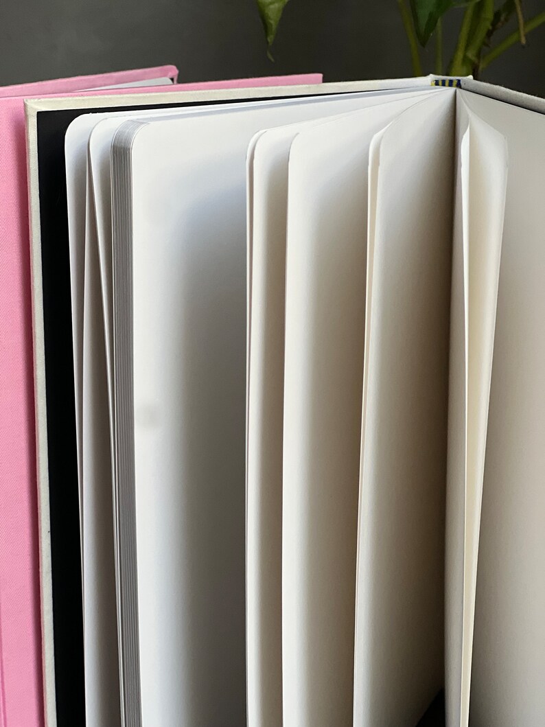 Op de afbeelding: Een open notitieboek met witte pagina's, dat het interieur onthult. De pagina's zijn uitgespreid, wat hun dikte en textuur laat zien. Links is een roze boek zichtbaar en een zwarte rug is zichtbaar achter de open pagina's.