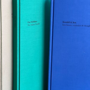 Op de afbeelding: Een verzameling hardcover boeken in verschillende kleuren. De boeken zijn in tinten beige, teal en blauw. Tekst op de ruggen en covers bevat "Our Holidays", "The Adams Family" en "Kendall & Ben love letters, reminders & thoughts."