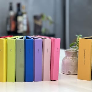 Op de afbeelding: Een verzameling kleurrijke hardcover boeken, waaronder rood, geel, groen, blauw en roze. Elke rug van het boek is gemarkeerd met "Year" en een nummer. Twee extra boeken zijn goud en groen, met namen en data.