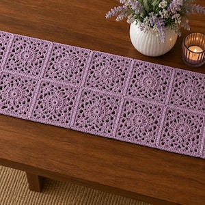 May include: A rectangular table runner made of twelve joined lavender crochet squares. Each square features a detailed circular floral pattern with openwork stitching. The runner is laid out flat, showcasing the uniform light purple yarn construction.
