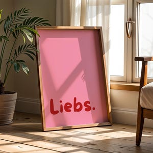 Könnte beinhalten: Ein rechteckiger Holzrahmen mit einem rosafarbenen Poster, auf dem das Wort "Liebs." in fetten, abgerundeten roten Buchstaben steht. Der Rahmen steht aufrecht auf einem Holzboden und zeigt ein schlichtes Design mit einfarbigem Hintergrund.