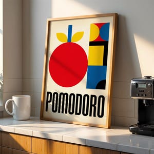 Könnte beinhalten: Ein gerahmter Kunstdruck mit einer roten Tomatenillustration und dem Wort "POMODORO" in fetten schwarzen Buchstaben. Der Druck wird auf einer Arbeitsplatte neben einer weißen Tasse und einer schwarzen Kaffeemaschine ausgestellt. Die Kunst hat einen modernen Stil.