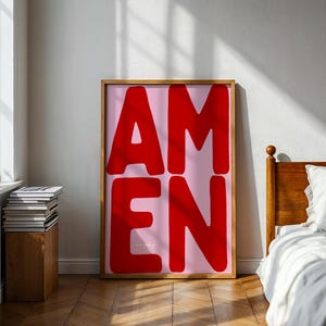 Könnte beinhalten: Ein rechteckiger Kunstdruck in einem hellen Holzrahmen mit dem Wort AMEN in großen, fetten, roten serifenlosen Großbuchstaben auf einem einfarbigen, hellrosa Hintergrund. Das Kunstwerk steht aufrecht auf einem Holzboden.