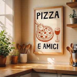 以下が含まれることがあります： 「PIZZA E AMICI」の文字が黒で書かれた額入りのプリント。ピザのイラストが描かれており、一切れが取り除かれ、ワイングラスが描かれています。画像は、植物や調理器具のあるキッチン環境にあります。