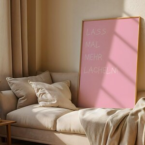 Könnte beinhalten: Ein rechteckiges Wandbild in einem dünnen goldenen Rahmen mit einem einfarbigen rosa Hintergrund und dem weißen handschriftlichen Text LASS MAL MEHR LACHELN!, der in vier Zeilen angeordnet ist. Das Bild steht auf einem neutralen Sofa.