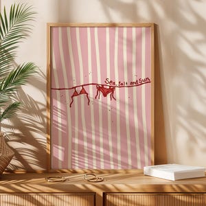 May include: A framed art print with a pink and white striped design. A red bikini is depicted hanging on a clothesline, with the words "Sea, Salt and Sun" above. The print is displayed on a wooden surface.