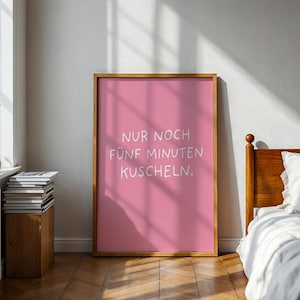 Op de afbeelding: Een roze ingelijste print met de tekst "NUR NOCH FÜNF MINUTEN KUSCHELN" in het wit. De print zit in een houten lijst en wordt weergegeven in een slaapkamer met een bed en een raam.