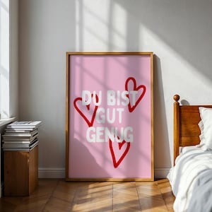 Peut inclure: Une affiche murale rectangulaire dans un cadre en bois présentant un fond rose clair avec trois contours de cœurs rouges dessinés à la main et le texte blanc "DU BIST GUT GENUG" centré sur l'impression, en orientation verticale.