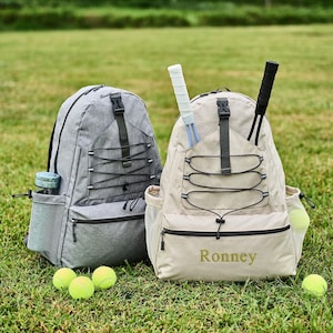Könnte beinhalten: Zwei Rucksäcke, einer grau und einer beige, stehen auf einer Wiese. Der beige Rucksack hat den Namen "Ronney" aufgestickt. Tennisschläger und Tennisbälle sind zu sehen. Eine graue Wasserflasche befindet sich in der Seitentasche des grauen Rucksacks.