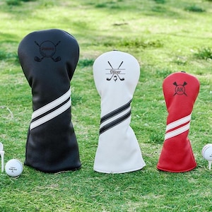 Könnte beinhalten: Drei Golfschlägerhauben in Schwarz, Weiß und Rot, jede mit einem einzigartigen Design und personalisiertem Text. Die Bezüge haben weiße Streifen und befinden sich auf einem grünen Rasen mit Golfbällen und Tees.