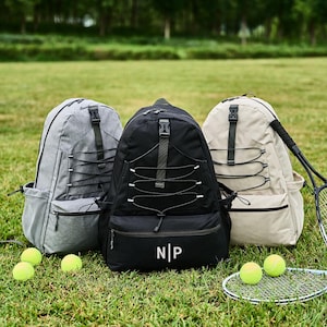 Può includere: Tre zaini grigi, neri e beige, con una racchetta e palline da tennis su un campo erboso. Lo zaino nero ha le iniziali "NP" sul davanti. È visibile anche una borraccia.