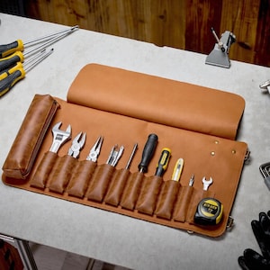 Peut inclure: Une trousse à outils en cuir beige dotée de multiples compartiments verticaux contenant divers outils à main, dont une clé, des pinces, des tournevis et un mètre ruban jaune, avec un grand rabat et des détails en métal.