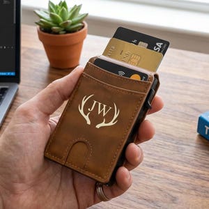 Op de afbeelding: Een bruine lederen kaarthouder met een in laser gegraveerd ontwerp van hertengeweien rondom de initialen JW. De portemonnee heeft een verticaal vak voor meerdere creditcards, waarbij drie kaarten gedeeltelijk uit de bovenkant steken.