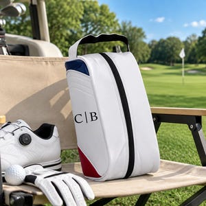 May include: A white golf shoe bag with a black zip, a top handle, and red and blue accents. The bag features the black monogram letters C|B printed on the front. The bag is positioned next to a white golf shoe and a white golf ball on a tee.