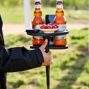May include: A black plastic outdoor drink holder with two circular cutouts for bottles, a central slot for a smartphone, and a small white bowl filled with red cherry tomatoes resting on the top surface between two amber glass beer bottles.