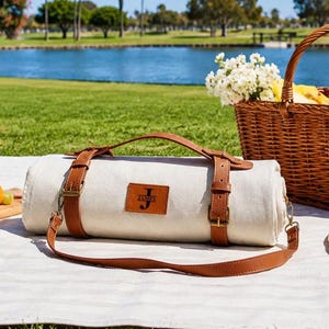May include: A rolled off-white canvas picnic blanket secured with brown leather straps and a matching shoulder carry strap. A rectangular brown leather patch is centered on the roll, featuring the letter J and the name JAMES in black text.