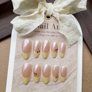 Puede incluir: Un conjunto de uñas postizas ovaladas con base rosa claro y puntas amarillas. Cada uña presenta un pequeño diseño de cereza. Las uñas se muestran en una tarjeta con el texto "Nail Art" y un mensaje de agradecimiento.