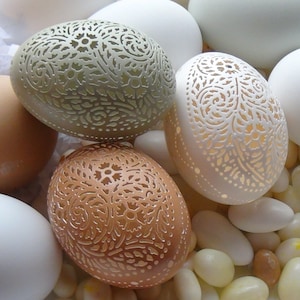 May include: Three intricately carved eggs with white, beige, and green patterns. The eggs are arranged on a surface with white and beige stones.