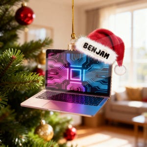 May include: A laptop-shaped Christmas ornament with a circuit board design in pink and blue. A Santa hat with the name "BENJAM" is on the ornament. The ornament is hanging in front of a Christmas tree.