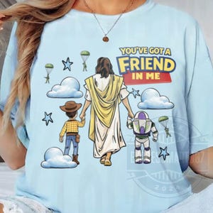 Può includere: Una t-shirt azzurra con una stampa grafica di una figura centrale in tunica gialla che tiene per mano un personaggio cowboy e un personaggio ranger spaziale. Il design include nuvole, stelle, soldati con paracadute e il testo YOU'VE GOT A FRIEND IN ME.