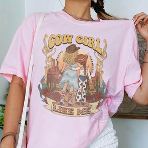 Puede incluir: Una camiseta de manga corta de color rosa claro con un estampado gr&aacute;fico de dos figuras con sombreros de vaquero en un paisaje des&eacute;rtico con cactus y rosas. El texto "COW GIRL" aparece arriba y "LIKE ME" en una cinta en la parte inferior.