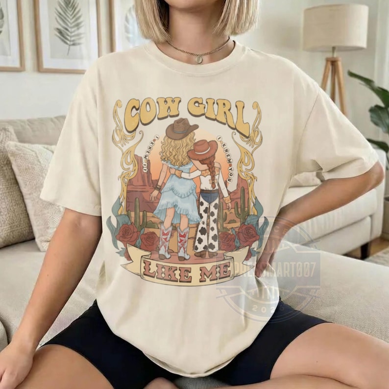 Puede incluir: Una camiseta de color crema con un estampado gr&aacute;fico de dos figuras con sombreros de vaquero en un paisaje des&eacute;rtico con cactus y rosas. El texto "COW GIRL" aparece en la parte superior y "LIKE ME" est&aacute; impreso en una cinta inferior.