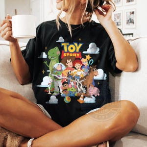 May include: A black short-sleeve t-shirt featuring a colorful graphic print of Toy Story characters including Woody, Buzz Lightyear, Jessie, Rex, and others, with the text Toy Story in yellow and red letters above the character group.