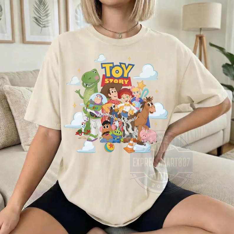 May include: A beige short-sleeve t-shirt featuring a colorful graphic print of Toy Story characters including Woody, Buzz Lightyear, Rex, Jessie, and Mr. Potato Head. The text TOY STORY is printed in bold blue and red letters above the characters.