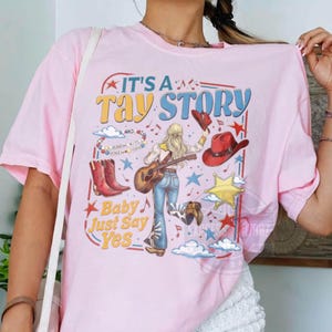 May include: A light pink t-shirt featuring a graphic print with the text "IT'S A TAY STORY" and "Baby Just Say Yes". The design includes a blonde figure playing an acoustic guitar, red cowboy boots, a red cowboy hat, stars, and clouds.