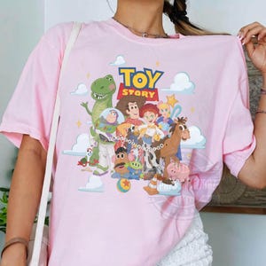 May include: A light pink t-shirt featuring a colorful graphic print of Toy Story characters including Woody, Buzz Lightyear, Rex, Jessie, and others, with the Toy Story logo at the top and white clouds in the background on a soft fabric base.