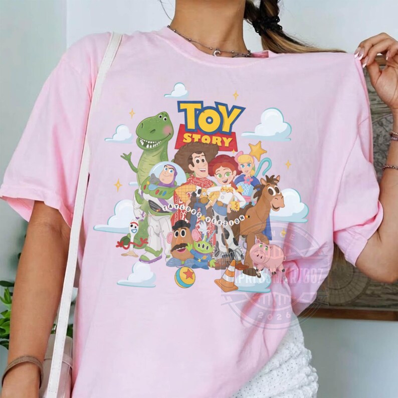 May include: A light pink t-shirt featuring a colorful graphic print of Toy Story characters including Woody, Buzz Lightyear, Rex, Jessie, and others, with the Toy Story logo at the top and white clouds in the background on a soft fabric base.