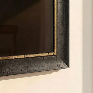 Puede incluir: Primer plano de una obra de arte enmarcada en negro con un borde interior dorado. El marco tiene un aspecto texturizado, similar a la madera. La superficie de la obra es oscura, reflejando el entorno. El marco está montado en una pared blanca.