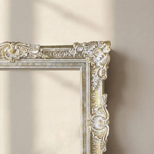 Puede incluir: Marco de fotos rectangular y ornamentado, con acabado dorado y blanco. El marco presenta diseños florales y de volutas esculpidos y detallados. El marco está vacío, con un fondo neutro visible a través de la abertura.